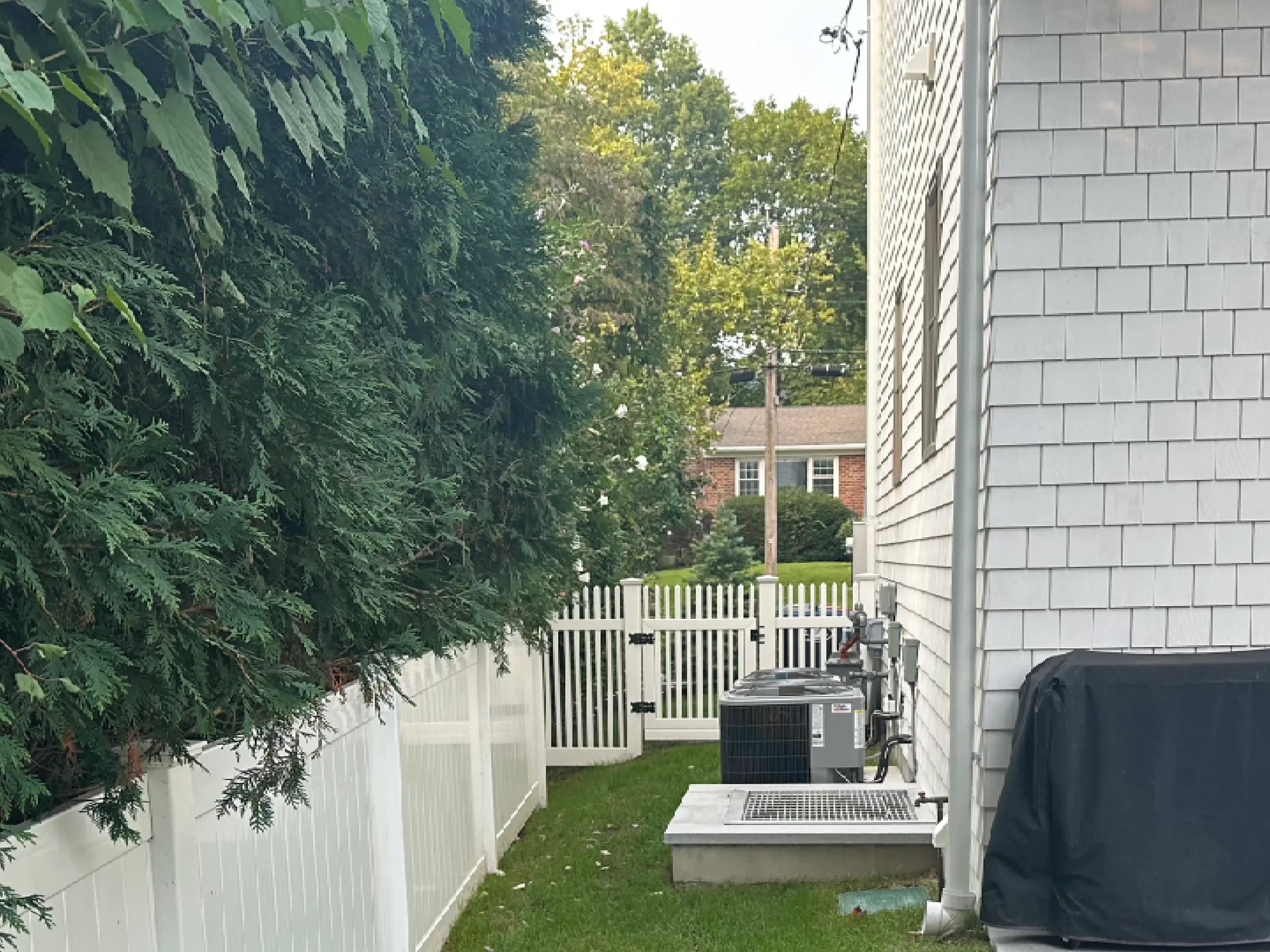 A narrow backyard with green grass, white fencing, and a brick house.